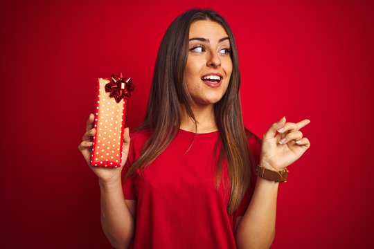 Young beautiful woman holding birthday gift standing over isolated red background very happy pointing with hand and finger to the side