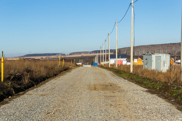 Fototapeta na wymiar Dirt road with electric poles and gas pipes in a cottage village. Autumn, sunny