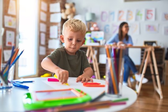 Young caucasian child playing at playschool with teacher. Mother and son at playroom drawing a draw with color pencils, young woman at the background sitting on desk.