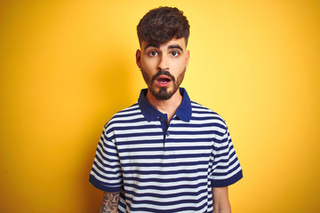 Young man with tattoo wearing striped polo standing over isolated yellow background afraid and shocked with surprise expression, fear and excited face.