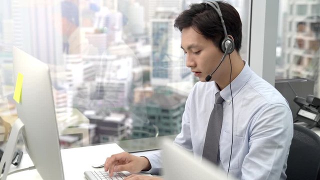 Young Asian man telemarketing agent wearing microphone headset typing on computer keyboard working in call center city office
