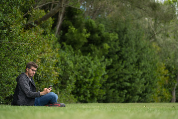 Naklejka na ściany i meble A young blond man is sitting with his legs crossed. He is carrying a laptop while activating Wi-Fi with his smartphone. He is in a beautiful and quiet park