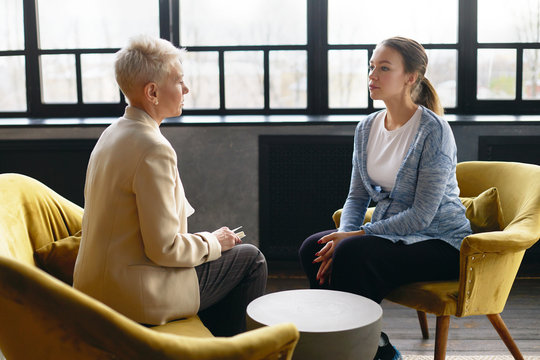 Side view image of two women sitting face to face in comfortable armchairs. Stylish mature female psychotherapist conducting counselling with young pregnant woman. Job interview and human resources