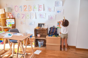 Beautiful toddler wearing glasses and unicorn diadem hanging up draw on the wall at kindergarten