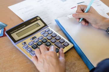 Hand of young woman using calculator to calculate the invoice and bill for monthly debt repay and writing down on empty paper on table at home office.