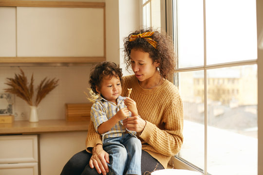 Pretty woman with curly hair sitting on windowsill with adorable baby on her lap, giving him toy or candy, little child looking with interest and curiosity. Motherhood, childcare and togetherness