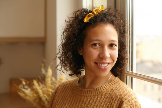 Indoor shot of charming friendly young casually dressed Latin woman with curly hair and brown skin posing by window in cozy kitchen interior, looking at camera with smile. Warm autumn colors