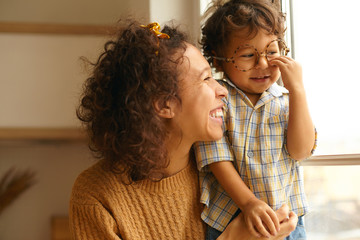 Close up image of happy young wavy haired Hispanic female posing by window embracing baby son. Cute...