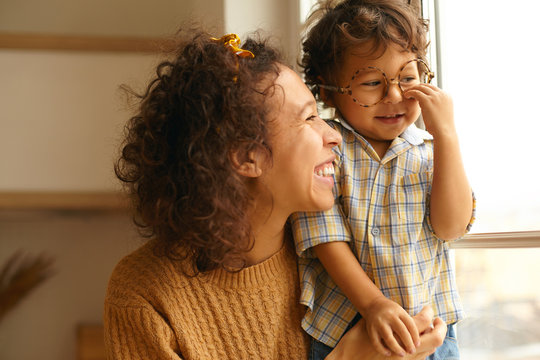 Close up image of happy young wavy haired Hispanic female posing by window embracing baby son. Cute three year old boy wearing round eyeglasses, spending day at home. Family and relationships