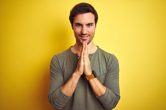 Young handsome man wearing casual t-shirt standing over isolated yellow background praying with hands together asking for forgiveness smiling confident.