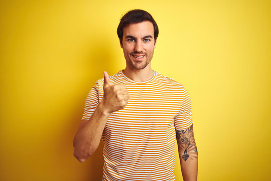Young handsome man with tattoo wearing striped t-shirt over isolated yellow background doing happy thumbs up gesture with hand. Approving expression looking at the camera showing success.