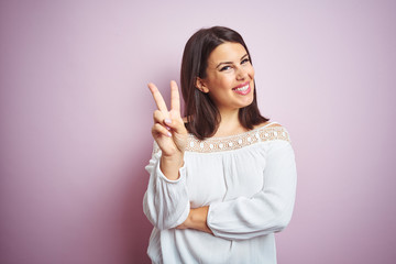 Young beautiful brunette woman over pink isolated background smiling with happy face winking at the camera doing victory sign. Number two.