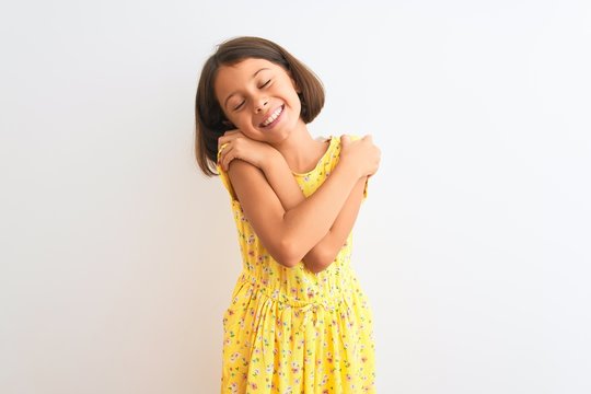 Young beautiful child girl wearing yellow floral dress standing over isolated white background Hugging oneself happy and positive, smiling confident. Self love and self care