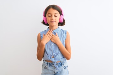 Beautiful child girl listening to music using headphones over isolated white background smiling with hands on chest with closed eyes and grateful gesture on face. Health concept.