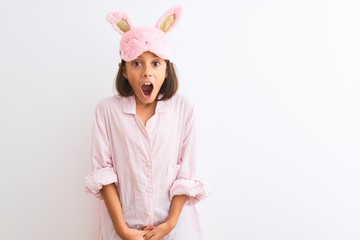 Beautiful child girl wearing sleep mask and pajama standing over isolated white background afraid and shocked with surprise expression, fear and excited face.