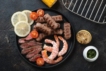 Surf and turf with beef meat, marbled beef sausages and shrimps, above view on a dark brown stone background, studio shot