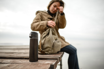 close-up of a thermos that stands on a village pier against the background of the silhouette of a girl buttoning a backpack