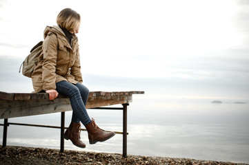 young girl in a warm jacket sits on the pier with his legs dangling and look towards the sea - 303759400