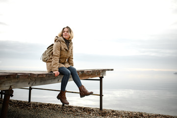 young girl in a warm jacket sits on a pier with legs dangling and look to the side