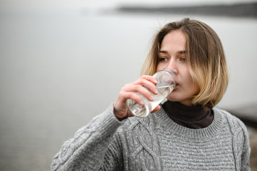 young girl in a warm woolen sweater drinks clean water from a reusable glass bottle by the sea