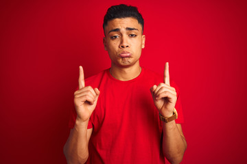 Young brazilian man wearing t-shirt standing over isolated red background Pointing up looking sad and upset, indicating direction with fingers, unhappy and depressed.