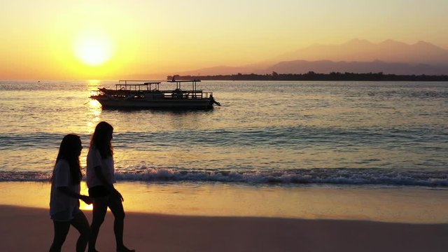 two teenage girls walking barefoot along hte sandy beach. tropical sunset over the ocean with island in the distance and boats in the water.