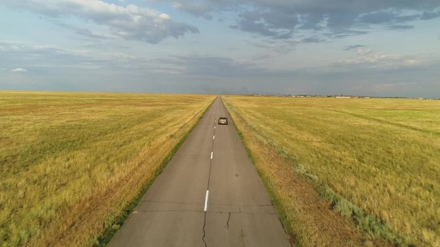 Drone follow up car travels at high speed leaves for horizon along picturesque asphalt straight road among deserted boundless steppe yellow field. Sunny clouds sunset. Trip freedom expanse. Break free