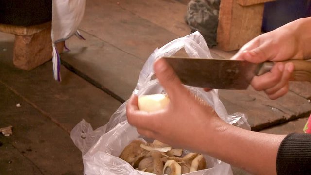 A daylight closeup shot of a local peeling and coring a potato from a plastic transparent bag using a medium-sized metal knife.