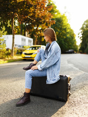 Profile portrait pensive girl sits on her suitcase on middle road, waiting a transportation, in sunset background. Horizontal view.