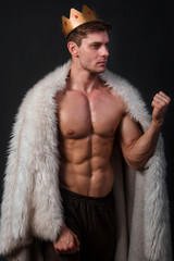 Portrait of a young bodybuilder with a crown on his head and a fur gown on his shoulders. Studio...