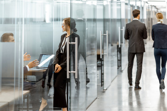 Business People Walking In The Hallway Of The Modern Office Building With Employees Working Behind Glass Partitions. Work In A Large Business Corporation