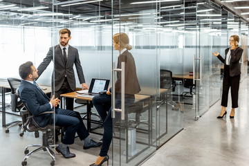 Group of strictly dressed business people having a discussion, sitting at the workplace with a computer in the modern office with glass partitions