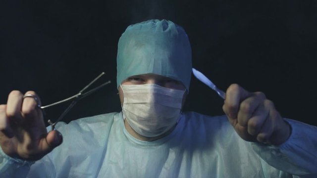 Doctor with forcep or grasper and scalpel looking at the camera and ready to start cut a patient's tissues in hospital. Surgery and operating table concept