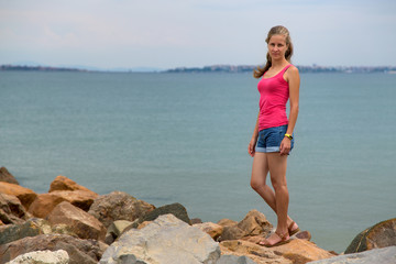 Young woman in summer clothes standing on big boulders on sea shore.