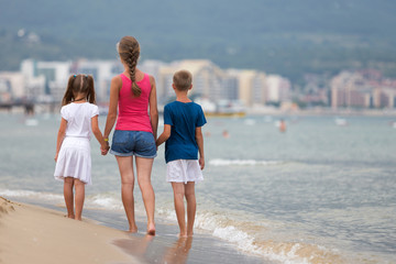 Mother and two children son and daughter walking together on sand beach in sea water in summer with bare feet in warm ocean waves.