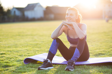 Young sportive woman in sports clothes sitting on training mat before doing exercises in field at...