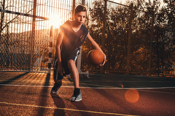Sports and basketball. A young teenager in a black tracksuit is playing a basketball. Sunset light....