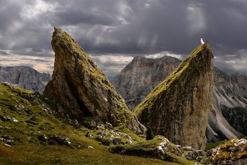 Woman standing on top of sharp rock looking at sunbeams through storm clouds in Dolomites. Concept of Faith and Hope.  Seceda ridge hike in Puez Odle Nature Park . Val Gardena. Italy.