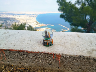 Glass snow globe stands on a white concrete surface against the background of seascape and cityscape of Antalya, Turkey. Concept of New Year and Christmas holidays in the south, winter without snow