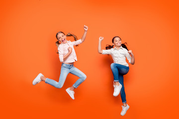 Full length photo of positive rejoicing two girlfriends jump rest relax win school sport competition raise fists wear white shirt denim jeans isolated over orange color background