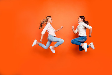 Full size profile side photo of positive funny two kids relax rest jump run play game wear white shirt denim jeans sneakers isolated orange color background