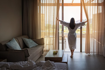 Woman opening curtains in hotel room in the morning - obrazy, fototapety, plakaty