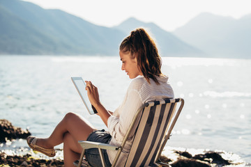 Smiling woman sitting on deck chair by the sea using tablet on a sunny day