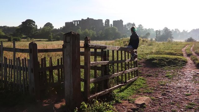 10 Year Old Boy Sitting On Gate Looking At Castle Ruins On Beautiful Early Summer Morning