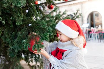 Cute child girl is decorating Christmas tree with red ornaments outside. Happy kid in santa hat at winter market on town street in Europe. Cozy fair and New Year in Dubrovnik, Croatia.