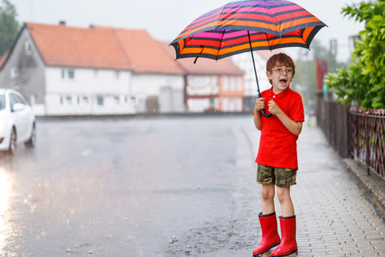 Kid boy wearing red rain boots and walking with colorful umbrella on city street. Child with glasses on summer day. happy kid during heavy summer shower rain.