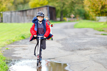cute little school kid boy riding on push scooter on the way to or from school. Schoolboy of 7 years driving through rain puddle. funny happy child in colorful fashion clothes and with helmet.