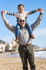 Happy senior man smiling joyfully piggybacking his cute little grandson at the beach