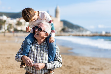 Happy senior man smiling joyfully piggybacking his cute little grandson at the beach - obrazy, fototapety, plakaty