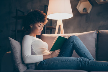 Photo of attractive dark skin curly lady holding favorite historic novel reading excited lying cozy couch wear casual sweater jeans outfit evening living room indoors